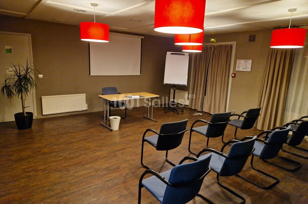 Salle de réunion avec chaises alignées, bureau, écran de projection, tableau blanc et éclairage par suspensions rouges.