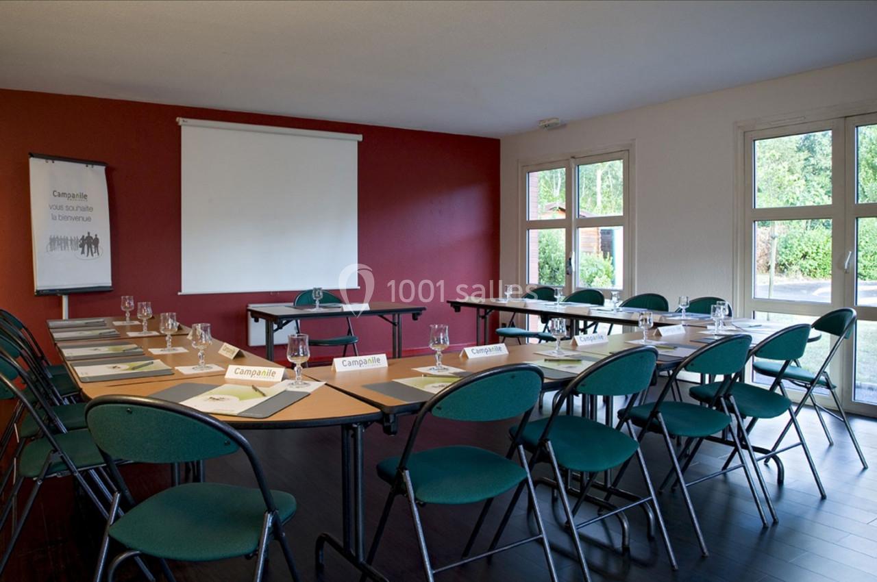 Salle de réunion lumineuse avec tables disposées en U, chaises vertes et écran de projection.