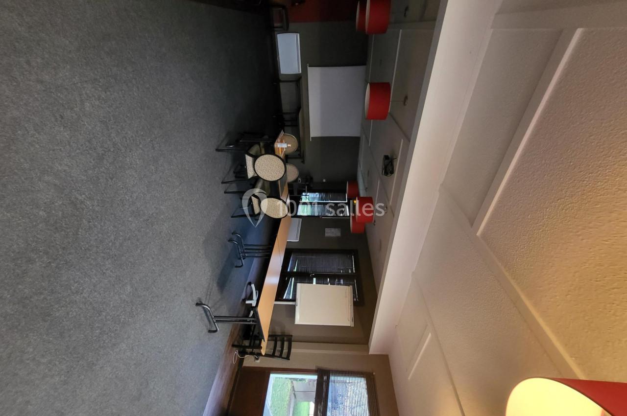 Salle de réunion vide avec chaises empilées, tables basses, écran de projection et éclairage suspendu.