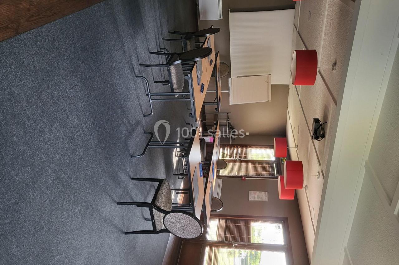Salle de réunion lumineuse avec tables en bois, chaises, écran de projection et suspensions rouges au plafond.