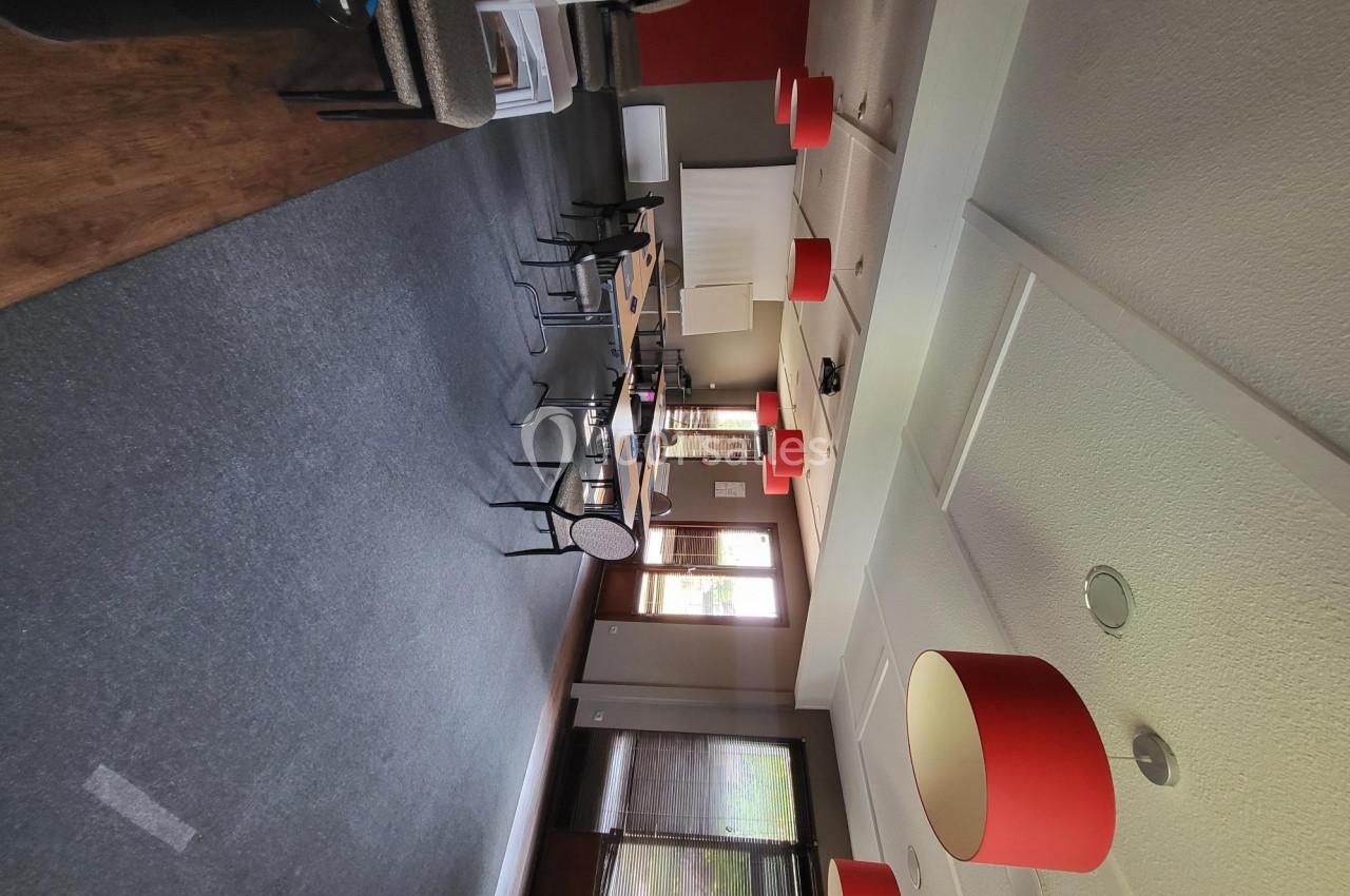 Salle de réunion vide avec tables, chaises, suspensions rouges et lumière naturelle provenant des fenêtres.