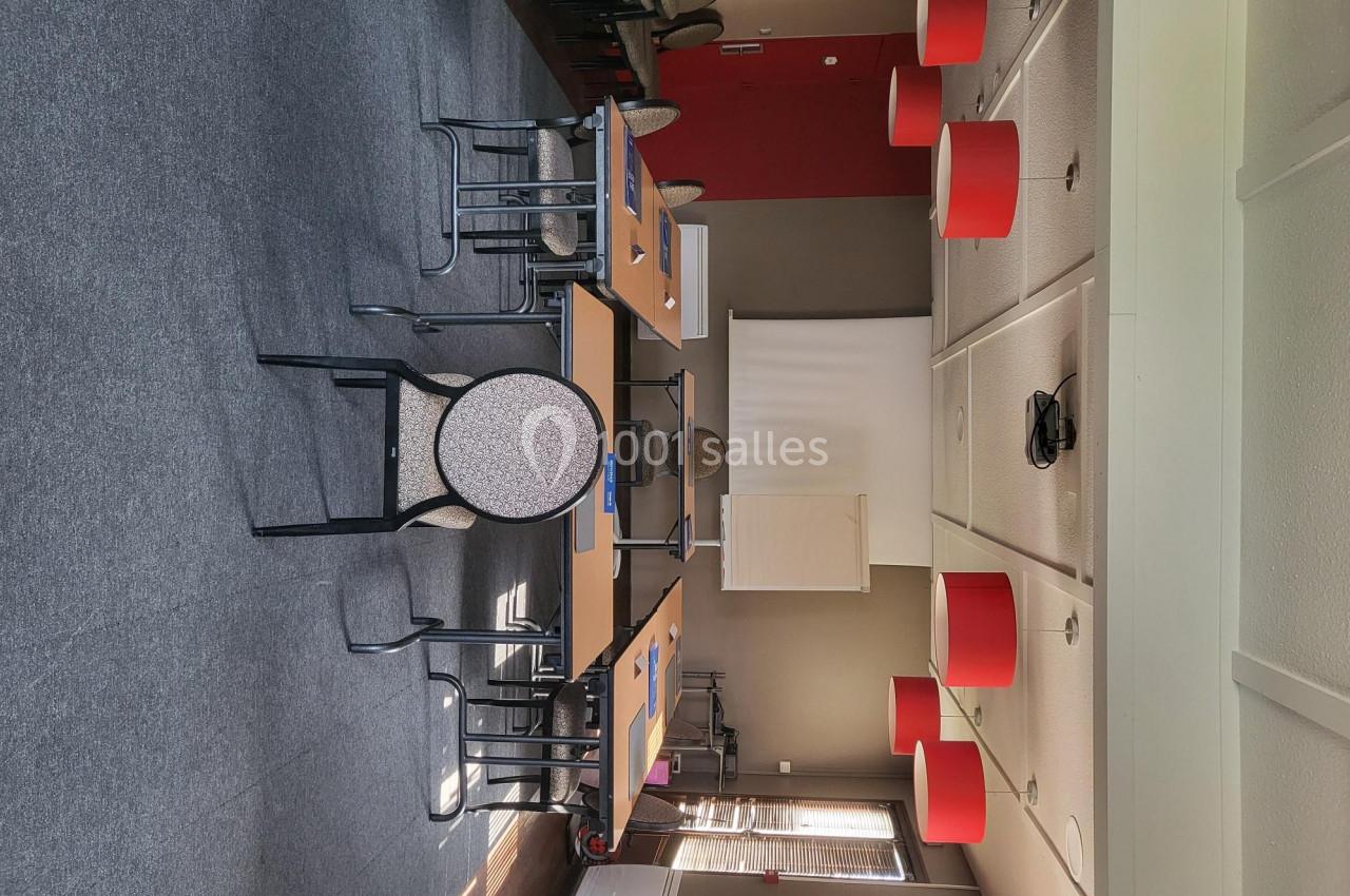 Salle de réunion avec tables disposées en U, chaises, écran de projection et luminaires rouges au plafond.