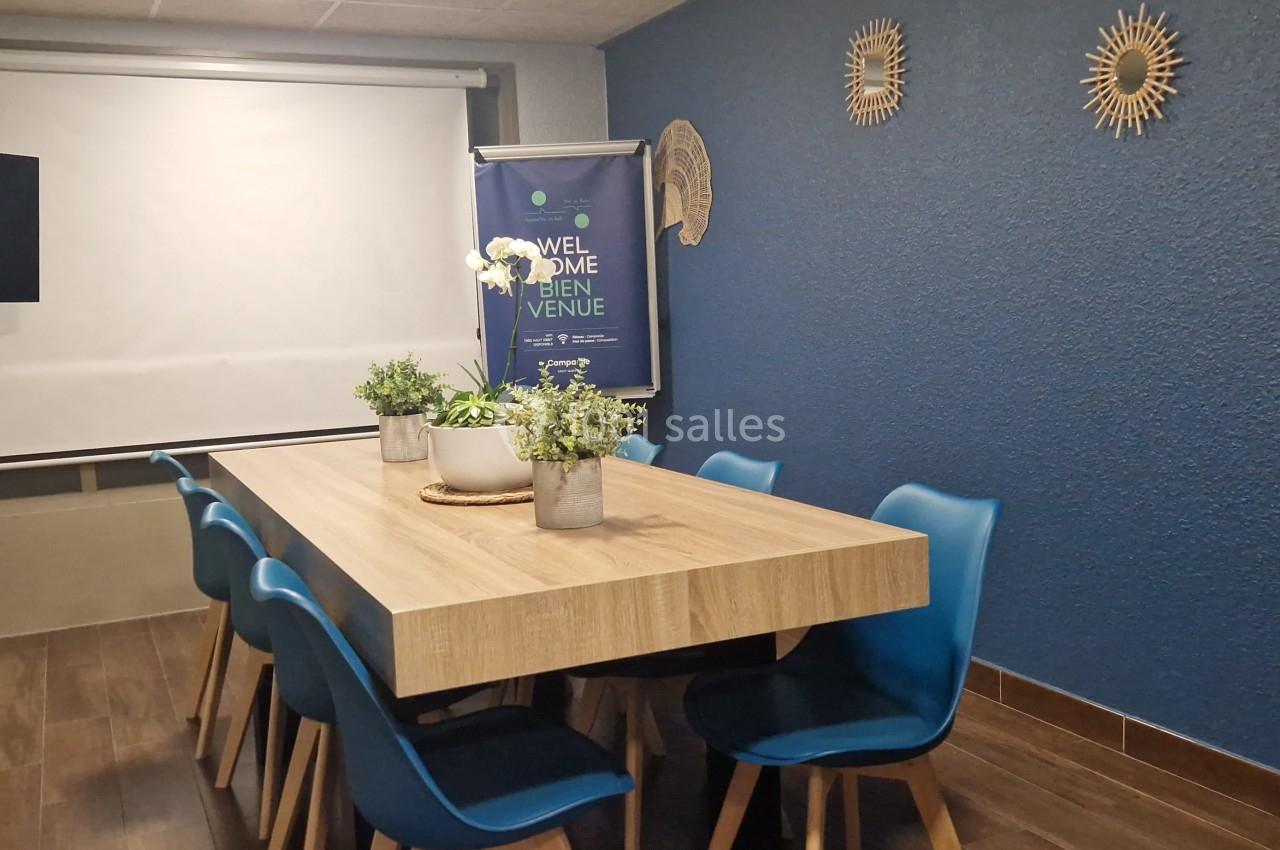 Salle de réunion avec table en bois, chaises bleues, plantes décoratives, écran de projection et mur bleu.