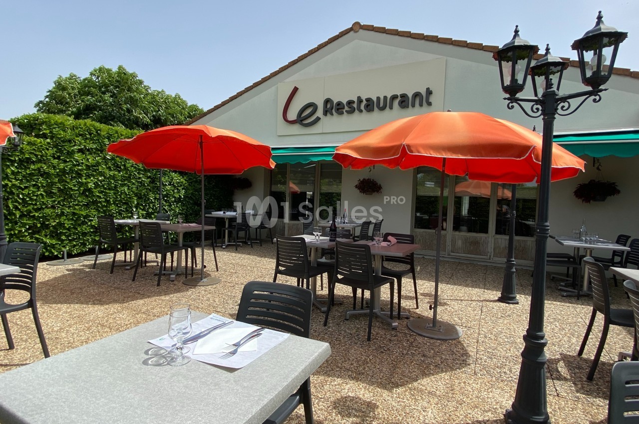 Terrasse d'un restaurant avec tables, chaises noires et parasols rouges, entourée de verdure par une journée ensoleillée.