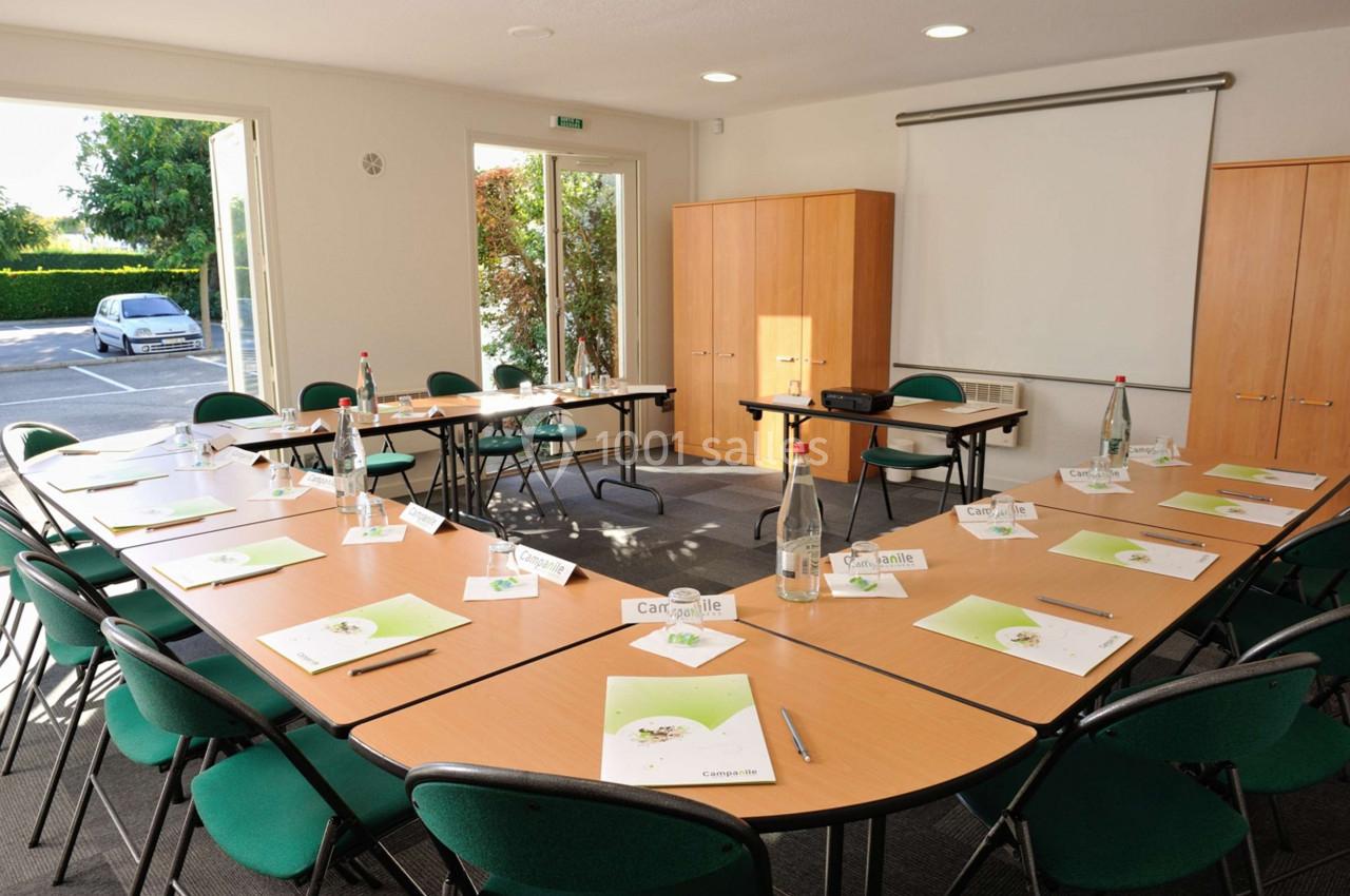 Salle de réunion lumineuse avec tables disposées en U, chaises vertes, documents, bouteilles d'eau et vidéoprojecteur.