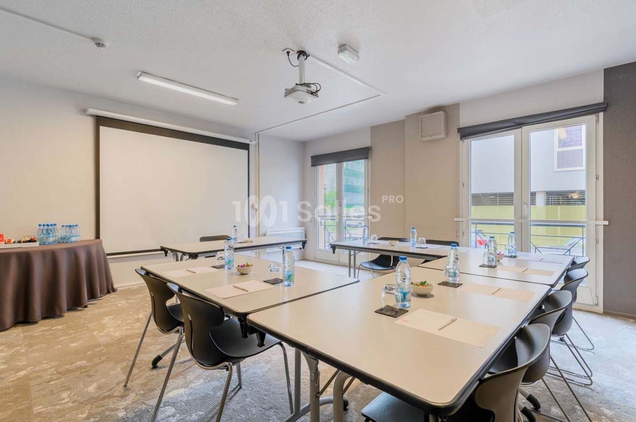 Salle de réunion lumineuse avec tables disposées en U, chaises, écran de projection et bouteilles d'eau.