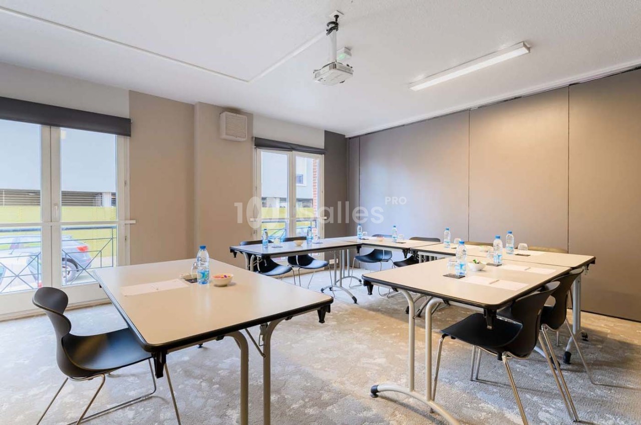 Salle de réunion lumineuse avec plusieurs tables, chaises noires, bouteilles d'eau et matériel de prise de notes.