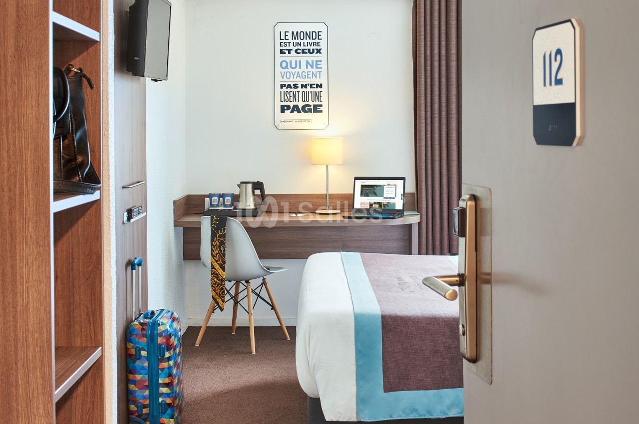 Chambre d'hôtel avec lit, bureau équipé d'un ordinateur portable, bouilloire, valise et décoration murale.