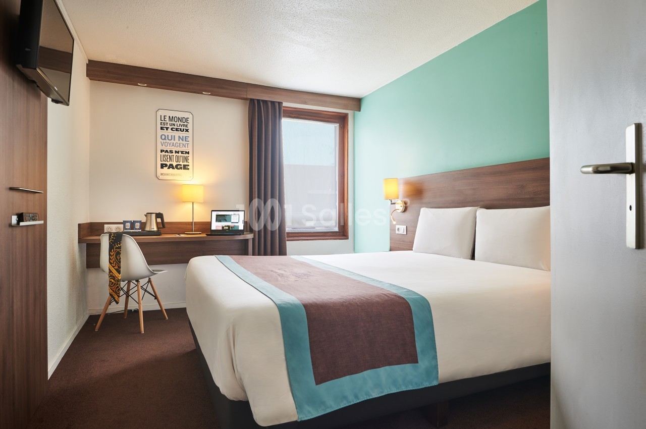 Chambre d'hôtel avec lit double, bureau, chaise, éclairage mural et mur turquoise, vue sur une fenêtre.