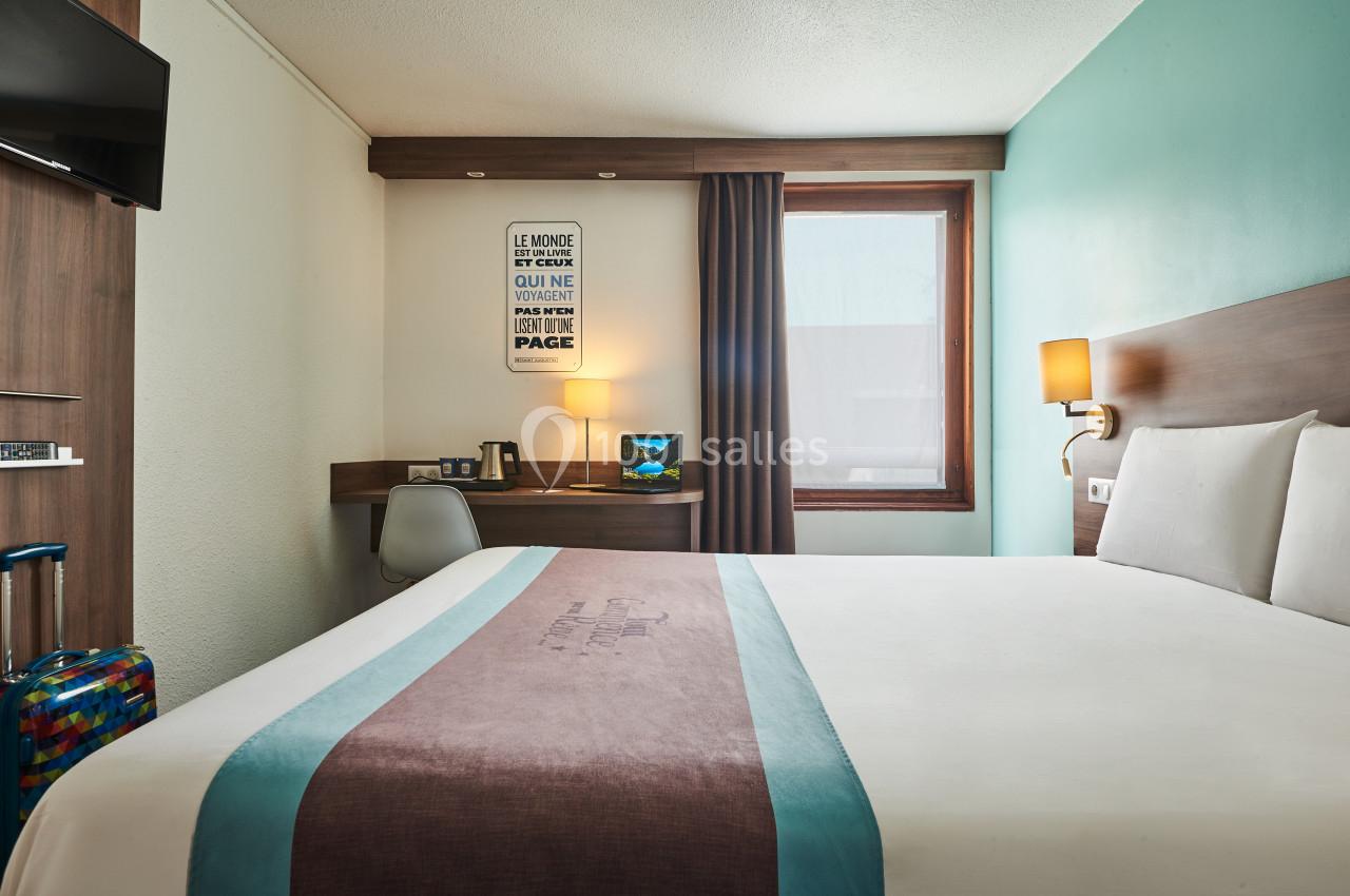 Chambre d'hôtel avec lit double, bureau, lampe allumée, valise colorée et fenêtre avec rideau partiellement ouvert.