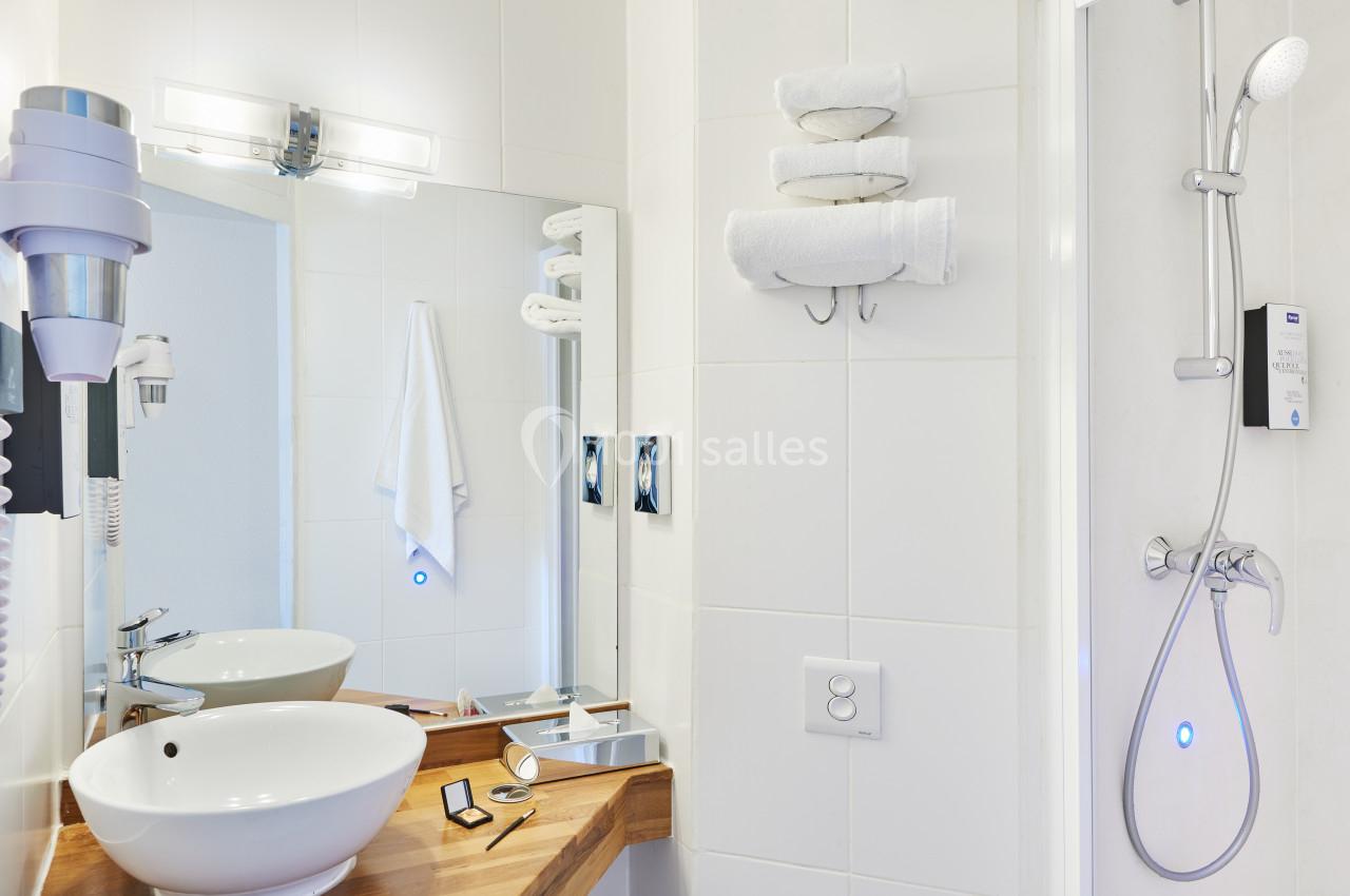 Salle de bain moderne avec lavabo, miroir, sèche-cheveux, serviettes pliées et douche équipée d'une pomme de douche.