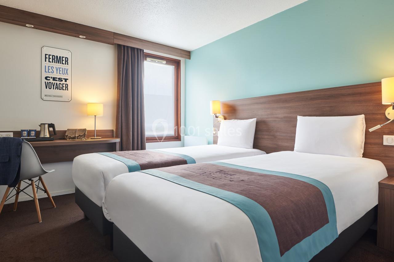 Chambre d'hôtel avec deux lits simples, murs bleu clair, bureau, chaise et fenêtre avec rideaux marron.