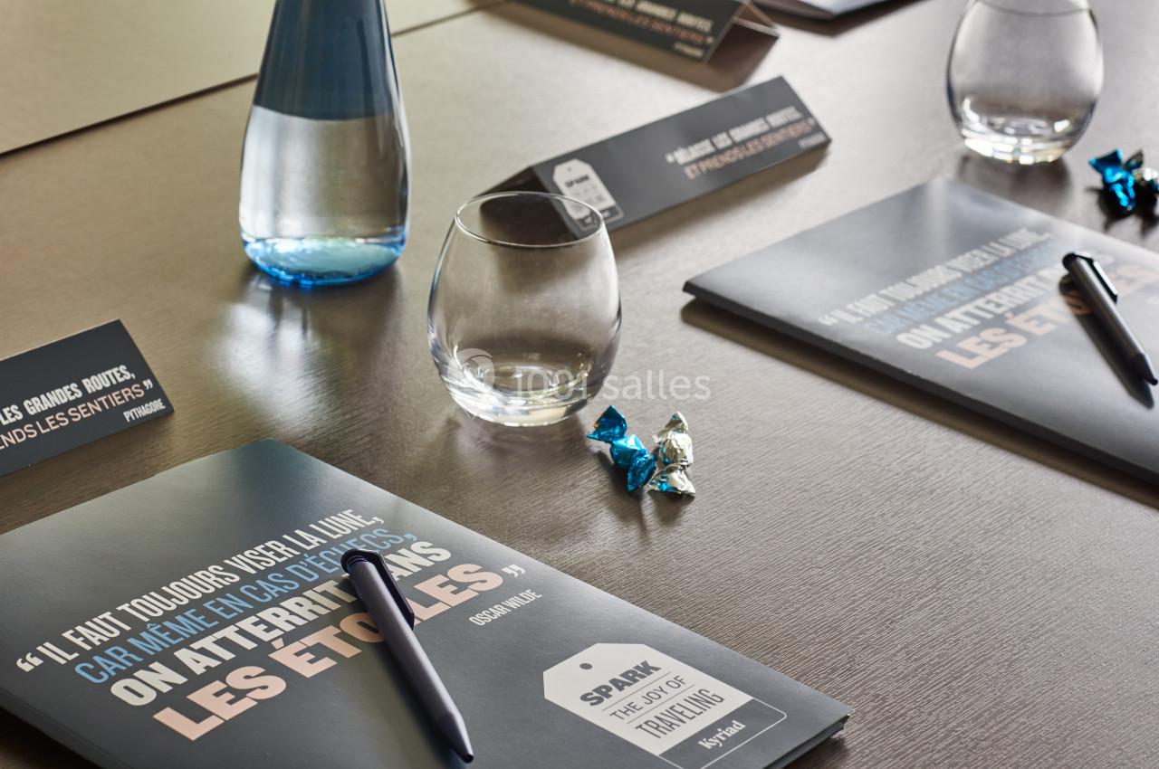 Bloc-notes, stylos, verres et bonbons disposés sur une table de réunion avec une bouteille d'eau en arrière-plan.