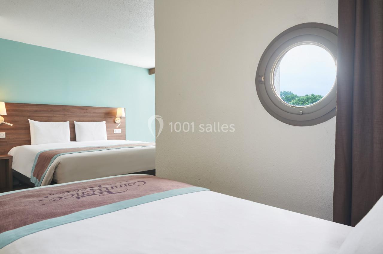 Deux lits dans une chambre d'hôtel lumineuse avec un hublot offrant une vue sur l'extérieur verdoyant.