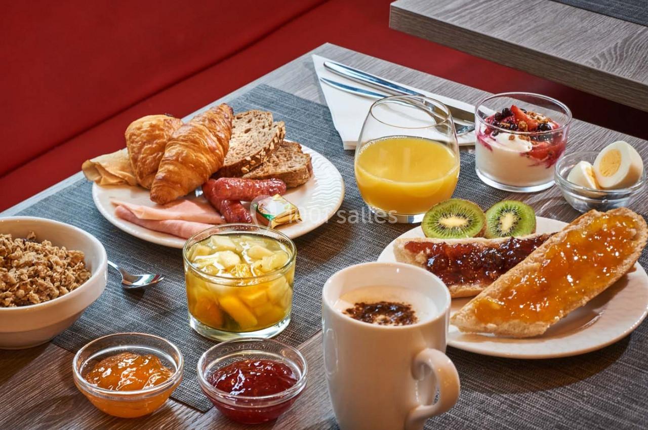 Petit-déjeuner varié avec viennoiseries, pain, confitures, fruits, yaourt, céréales, jus d'orange et café sur une table.