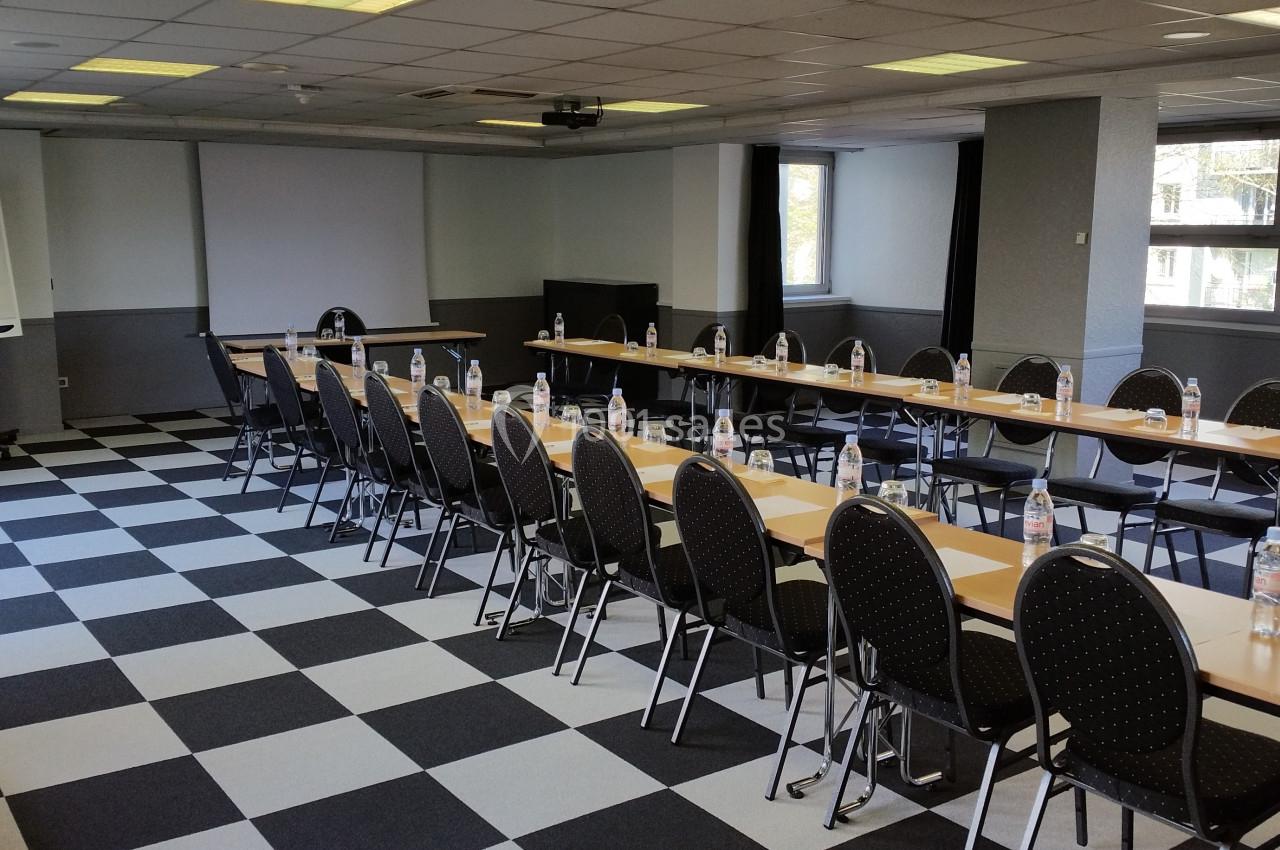 Salle de réunion lumineuse avec tables disposées en U, chaises noires, bouteilles d'eau et tableau de conférence.
