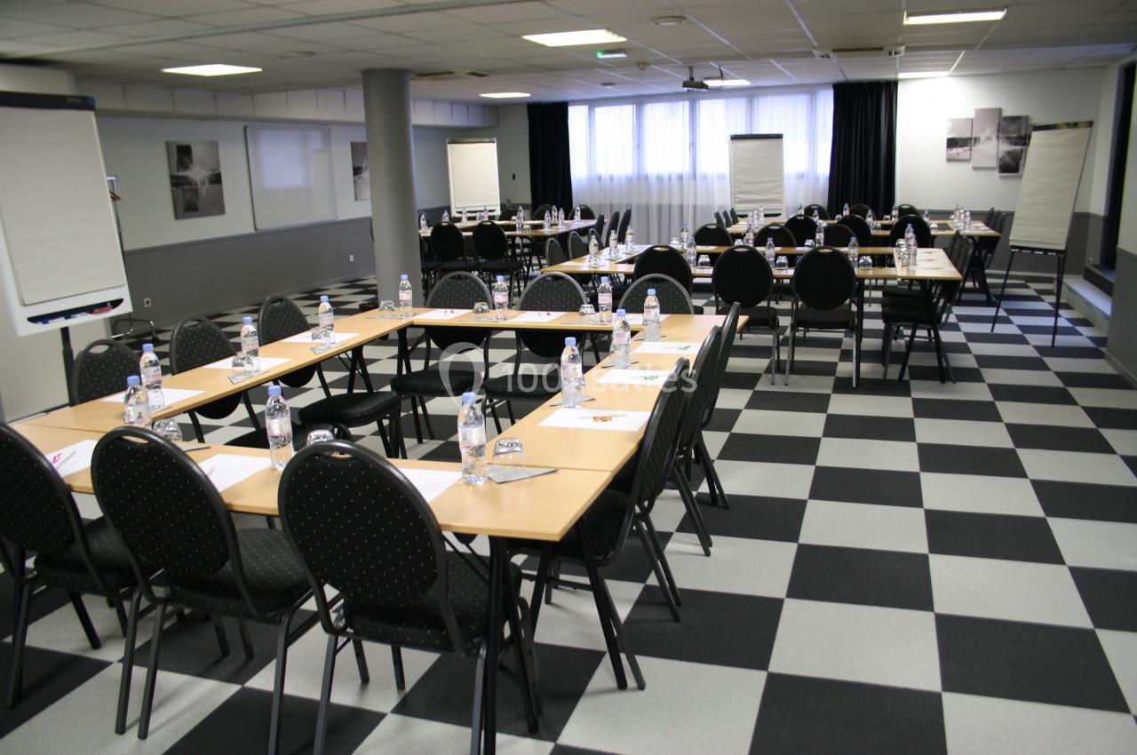 Salle de réunion équipée de tables disposées en U, chaises noires, bouteilles d'eau et chevalets de conférence.