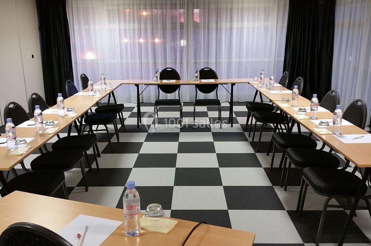Salle de réunion avec tables disposées en U, chaises noires, bouteilles d'eau et blocs-notes sur un sol à carreaux.