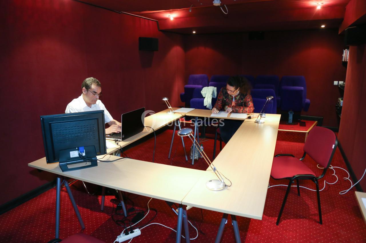 Deux personnes travaillent dans une salle équipée de tables en U, chaises, ordinateurs et éclairage tamisé.