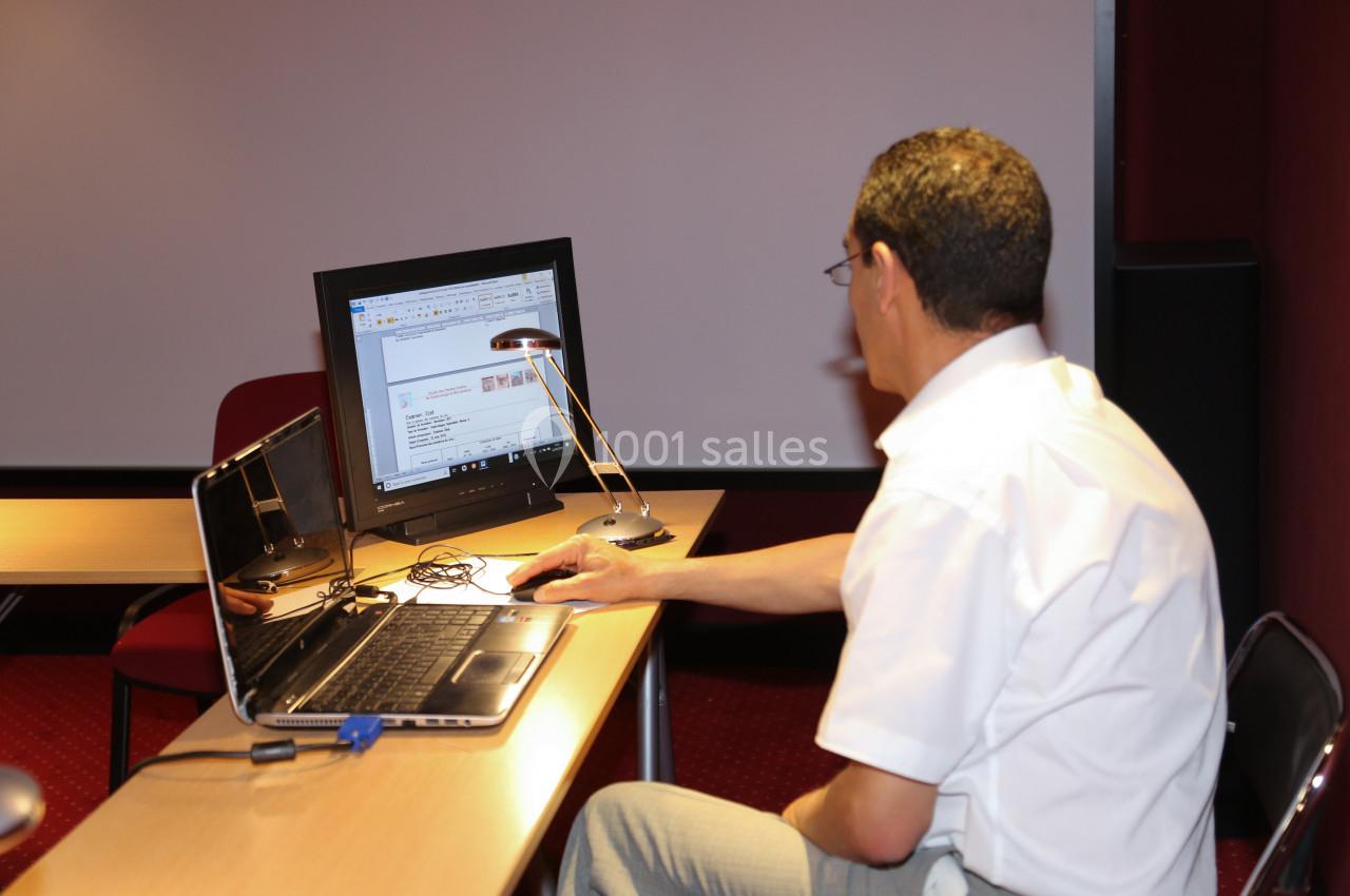 Un homme travaille sur un ordinateur portable et un écran externe dans une salle équipée d'un projecteur.