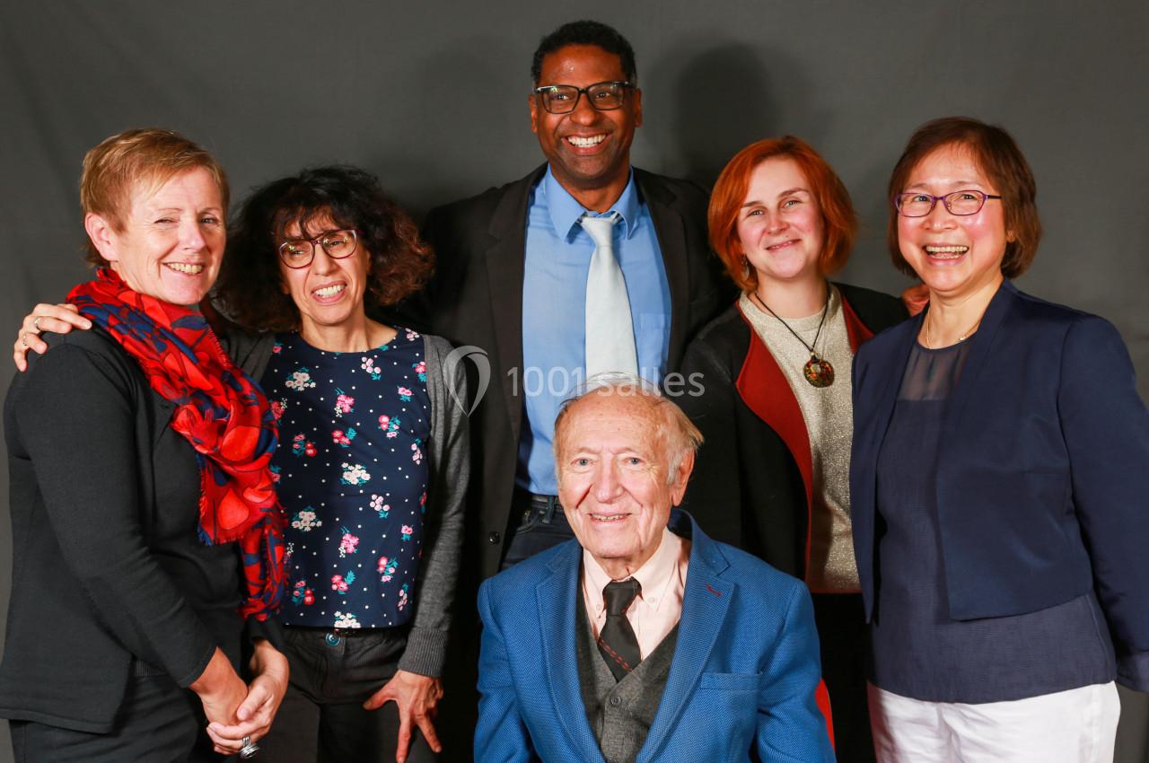 Un groupe de six personnes souriantes posant devant un fond gris, dont un homme âgé assis au centre.