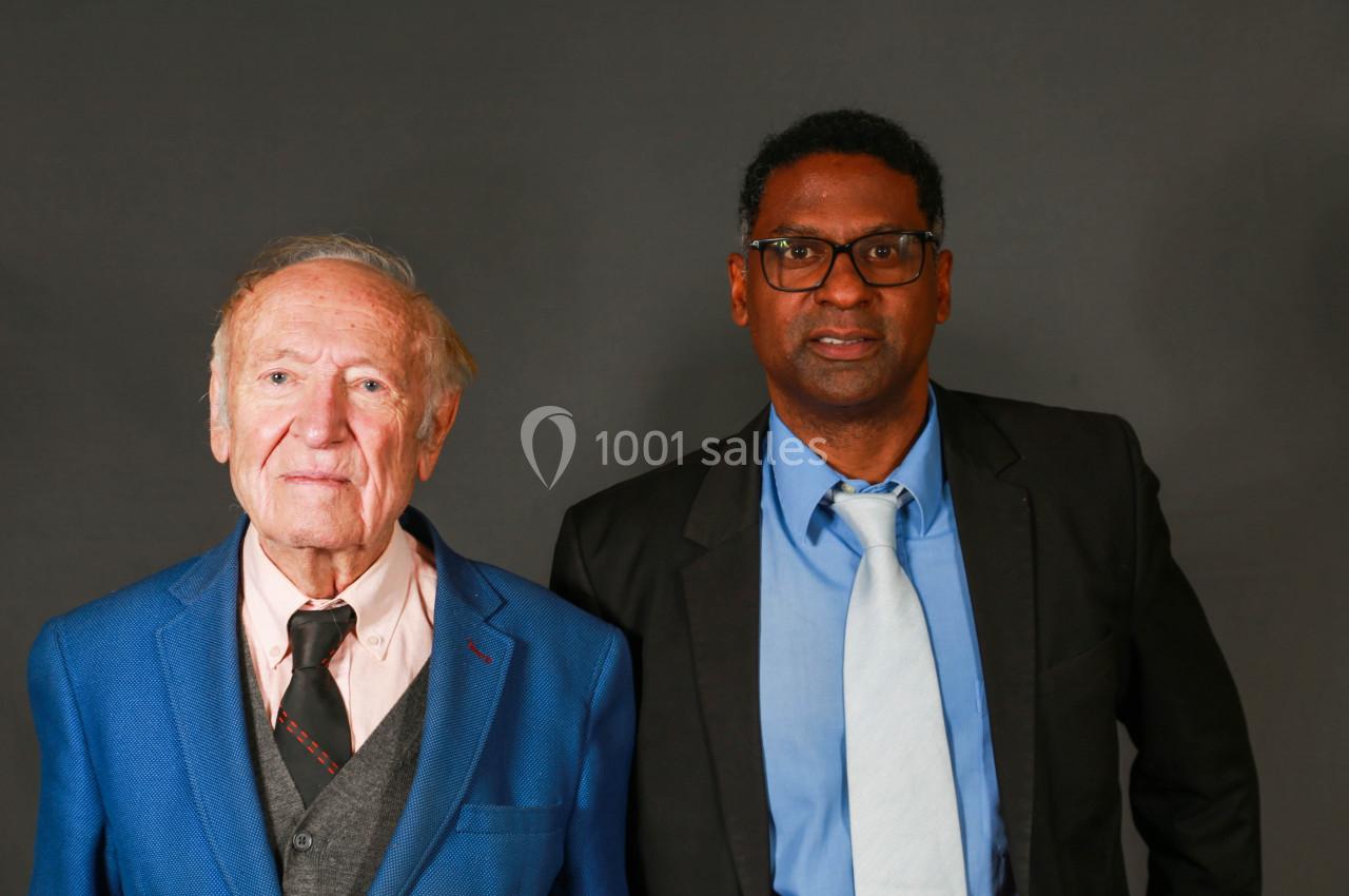Deux hommes en costume, l'un âgé et l'autre plus jeune, posent devant un fond gris uni.