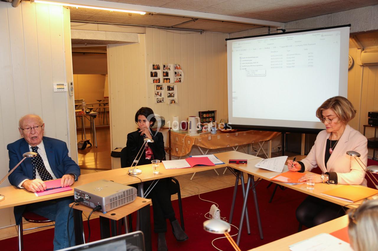 Trois personnes participent à une réunion autour de tables, avec un projecteur et un écran affichant un document.