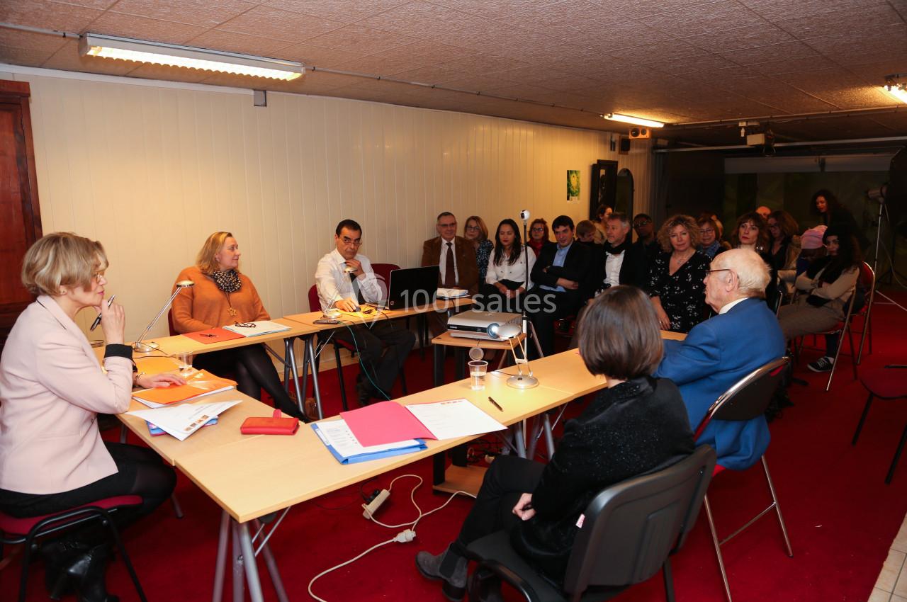 Un groupe de personnes assises dans une salle de réunion, écoutant une présentation autour de tables disposées en U.