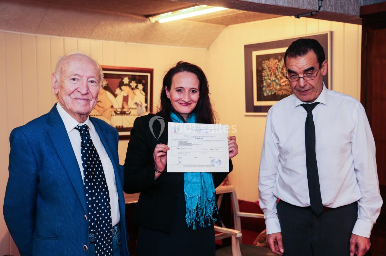 Une femme souriante tient un certificat entre deux hommes dans une pièce décorée de tableaux.