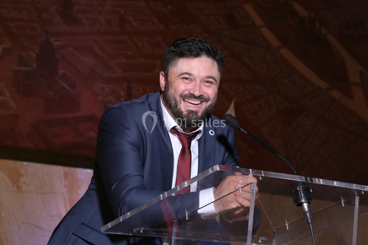 Homme souriant en costume et cravate rouge, debout derrière un pupitre transparent avec un micro, fond sombre.