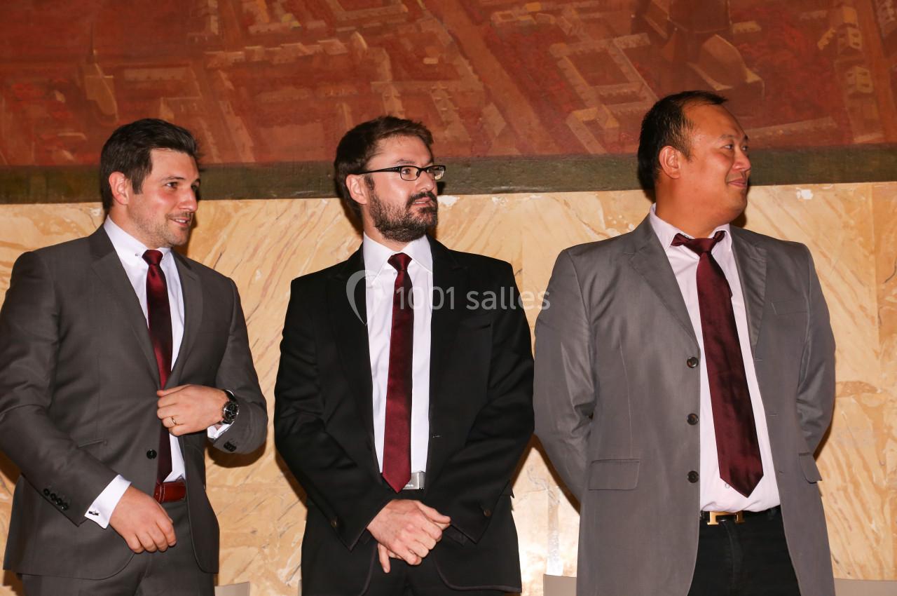 Trois hommes en costume et cravate bordeaux debout devant un mur beige et une fresque murale.