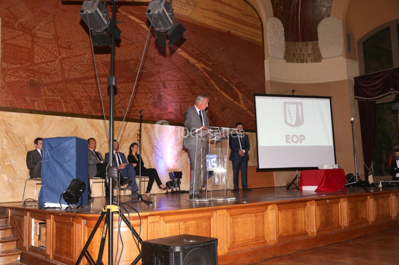 Un homme en costume prononce un discours sur une scène, devant un public, avec un écran projetant un logo en arrière-plan.