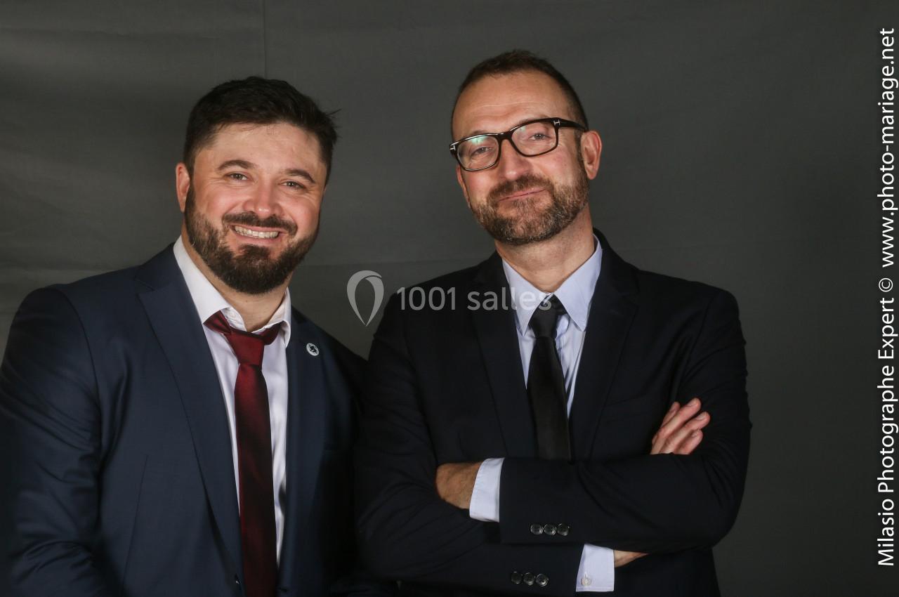 Deux hommes en costume posent côte à côte devant un fond gris, l'un souriant et l'autre avec les bras croisés.