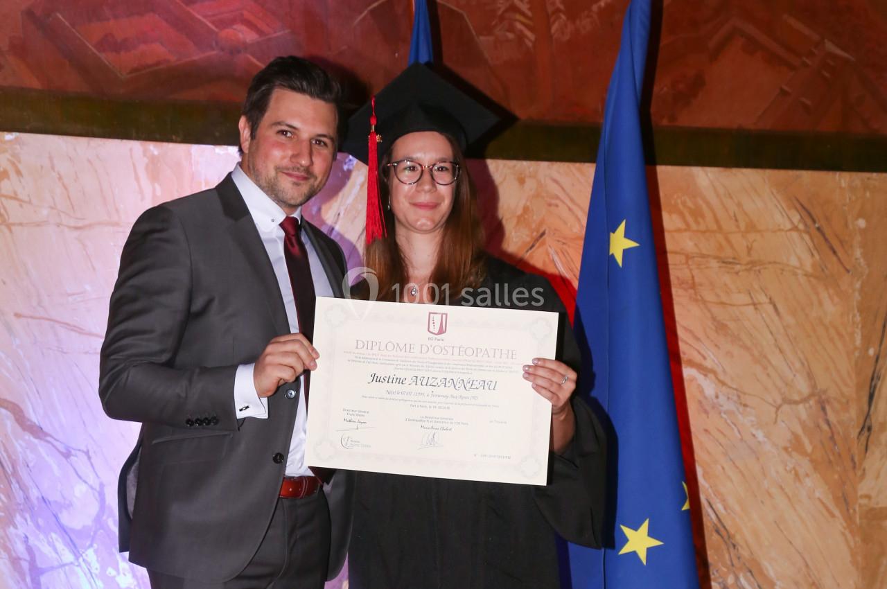 Une femme en tenue de diplômée tient un diplôme aux côtés d'un homme en costume, devant des drapeaux européens.
