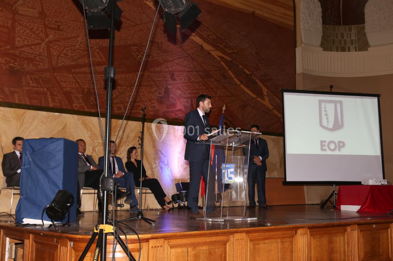 Un homme en costume parle debout à un pupitre transparent dans une salle avec écran de projection et public assis.