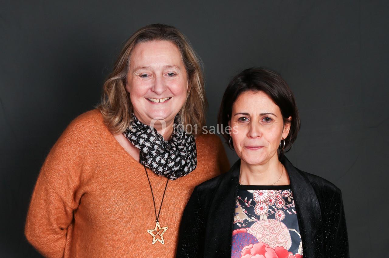 Deux femmes posant côte à côte devant un fond sombre, l'une portant un pull orange et l'autre une veste noire.