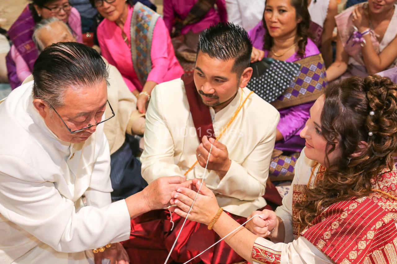 Un homme âgé attache un fil blanc aux poignets d'un couple en tenue traditionnelle, entouré de spectateurs.