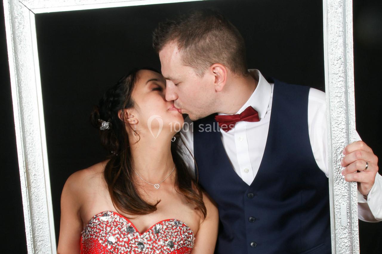 Un couple s'embrasse en tenant un cadre argenté, la femme porte une robe rouge scintillante et l'homme un costume avec nœud…