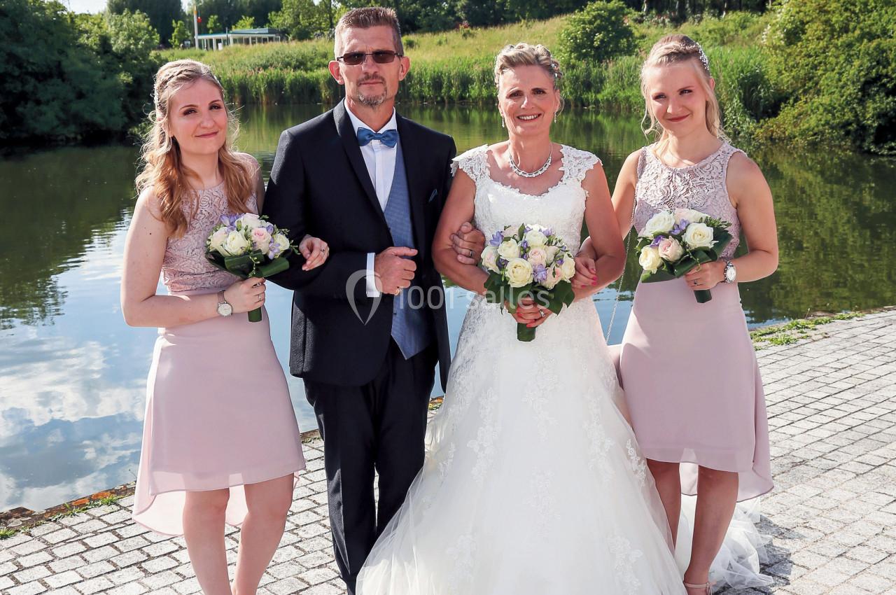 Un couple en tenue de mariage pose avec deux jeunes femmes tenant des bouquets, près d'un plan d'eau.