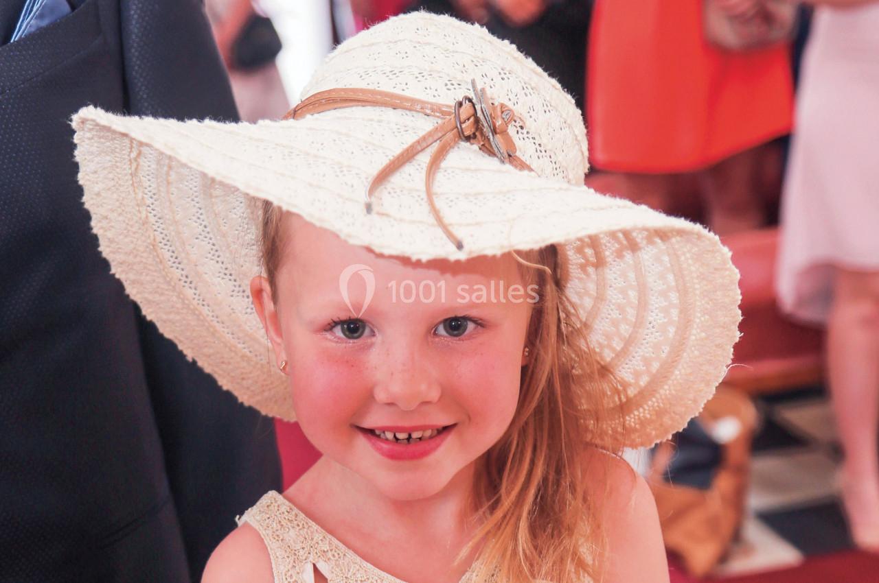 Une fillette souriante portant un chapeau en dentelle blanche dans un environnement intérieur animé.
