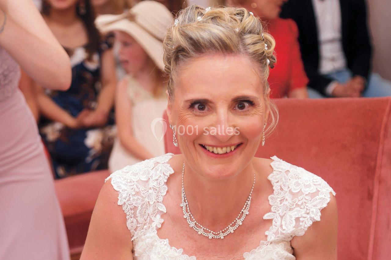 Femme souriante en robe blanche ornée de dentelle, assise sur un fauteuil rose, entourée de personnes en arrière-plan flou.