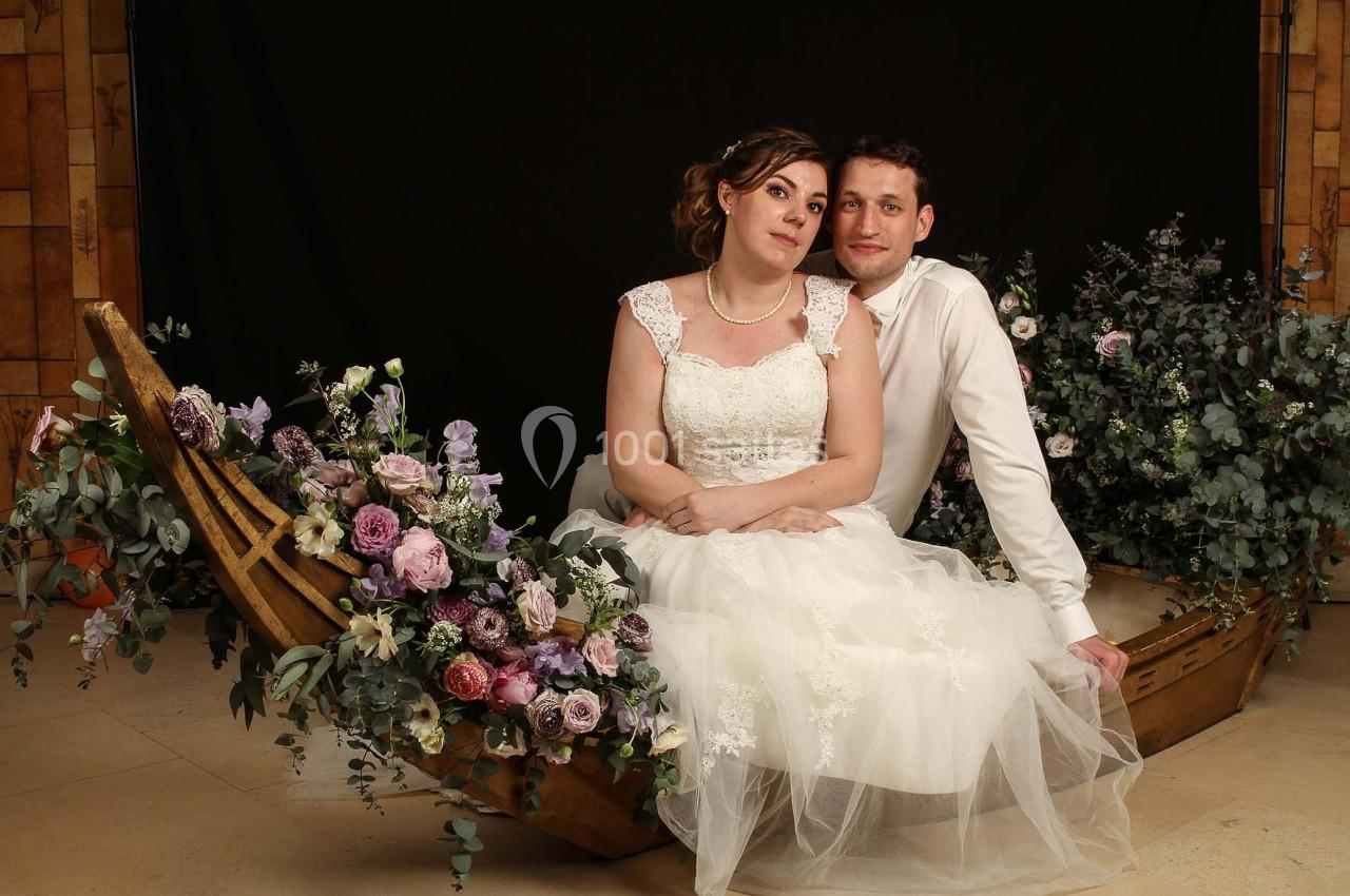 Un couple en tenue de mariage est assis dans une décoration florale en forme de barque, sur fond sombre.