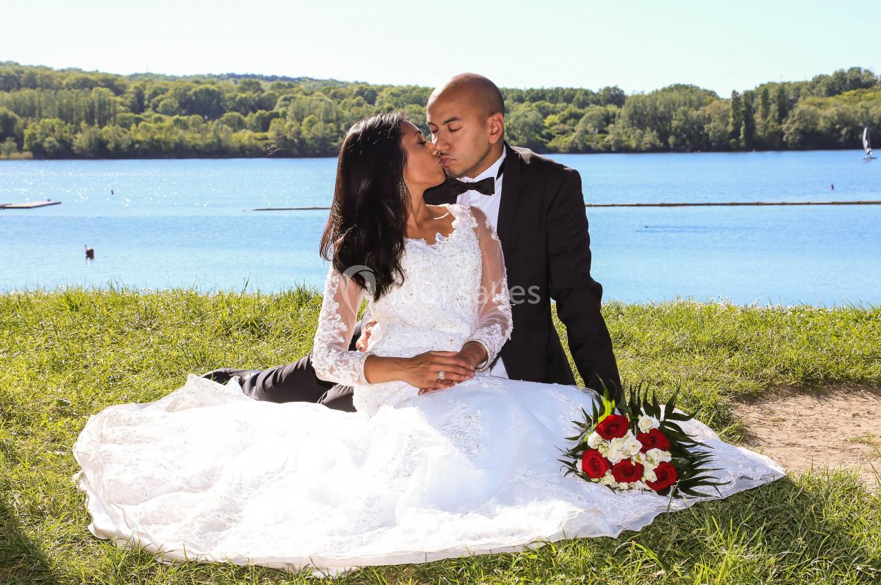 Un couple en tenue de mariage s'embrasse assis sur l'herbe près d'un lac, un bouquet de fleurs posé à côté.