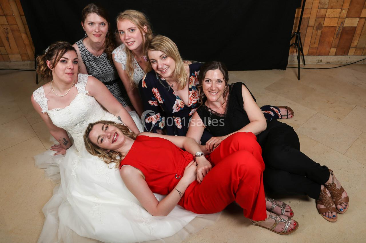 Six femmes souriantes posent ensemble, dont une en robe de mariée, assises ou allongées sur un sol clair.