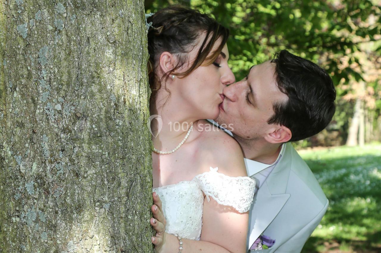 Un couple en tenue de mariage s'embrasse tendrement derrière un arbre dans un parc verdoyant.