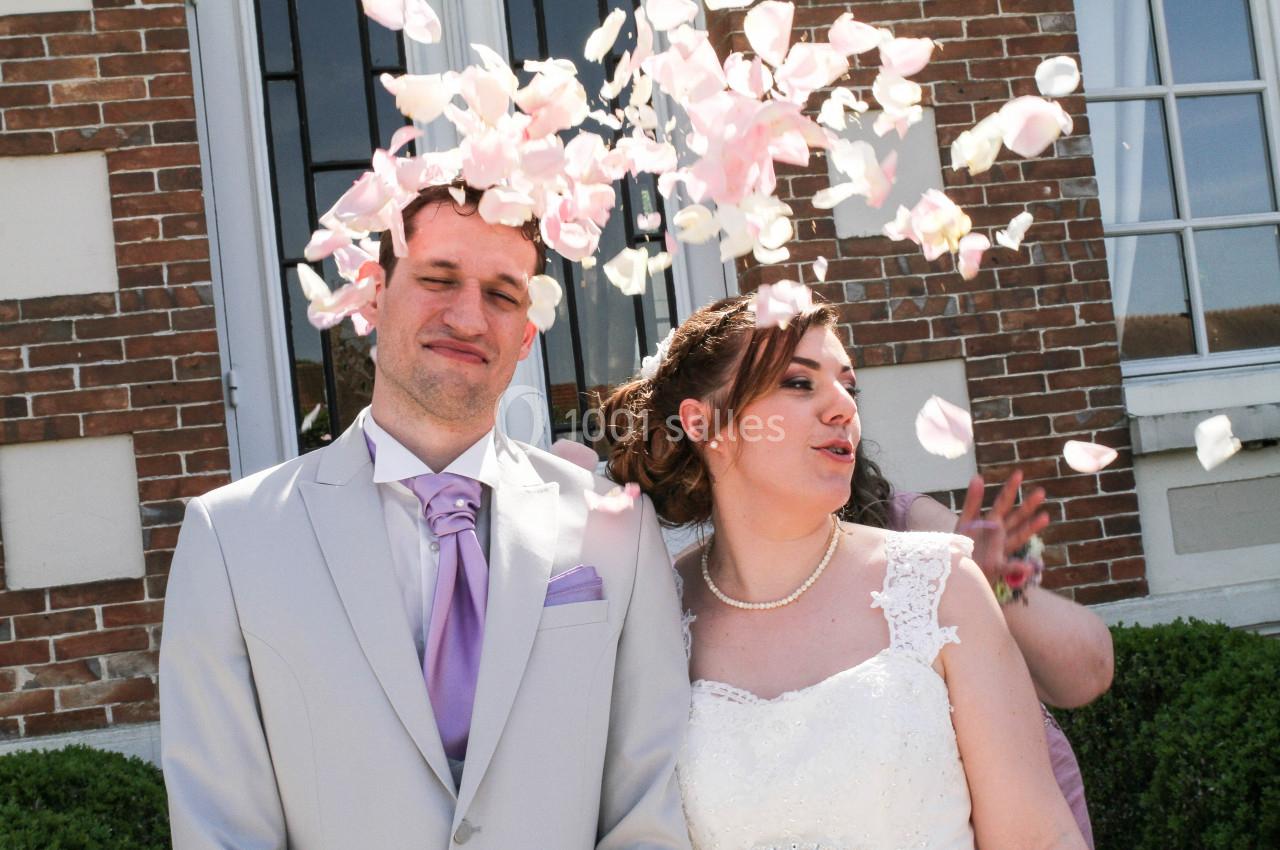 Un couple en tenue de mariage sourit sous une pluie de pétales devant un bâtiment en briques.