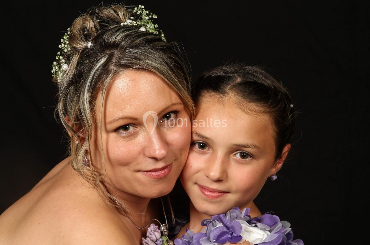 Une femme souriante et une jeune fille posent ensemble, vêtues de tenues ornées de fleurs violettes, sur fond noir.