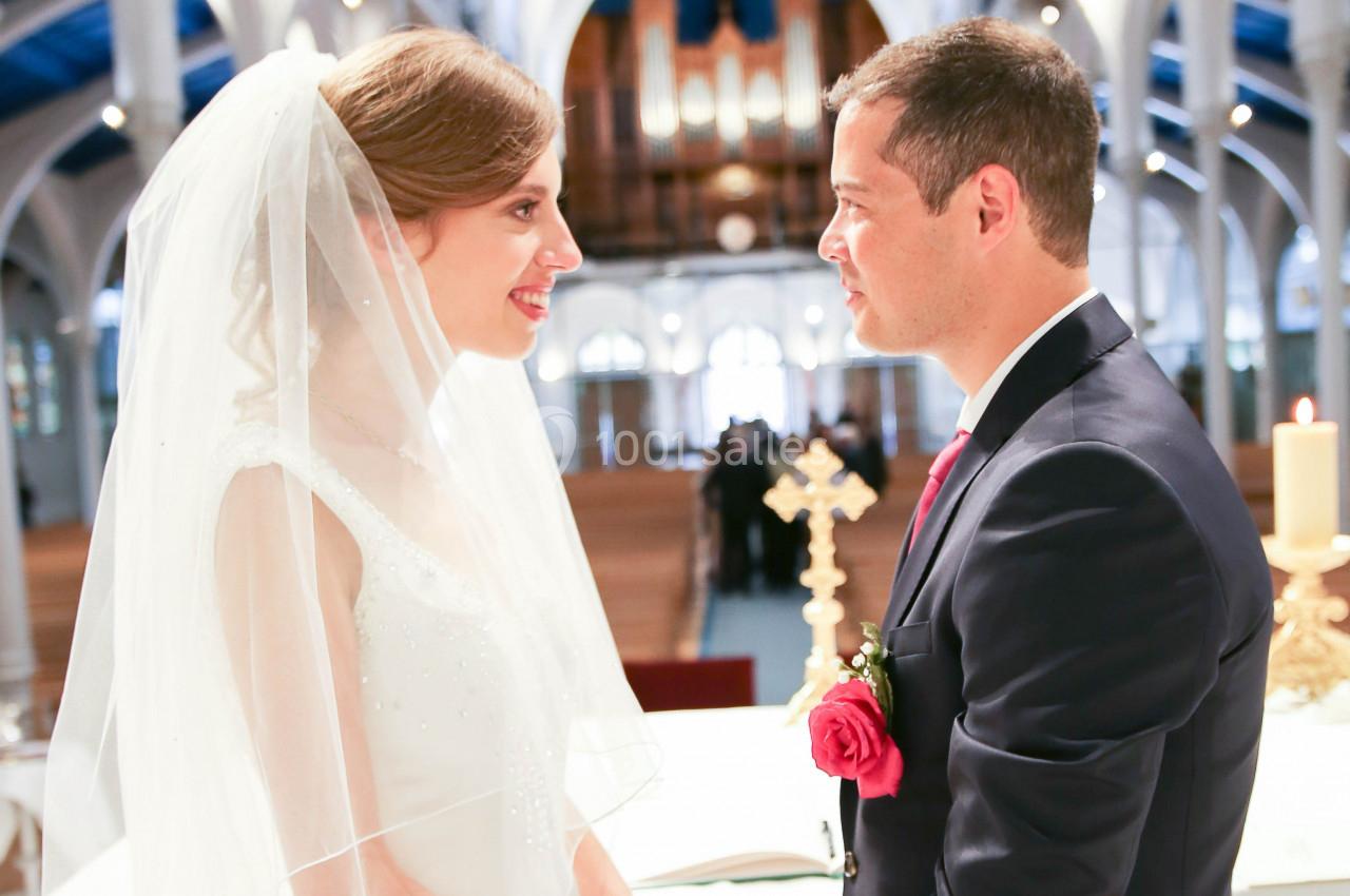 Un couple en tenue de mariage se regarde dans une église lumineuse, avec un autel et des bancs en arrière-plan.