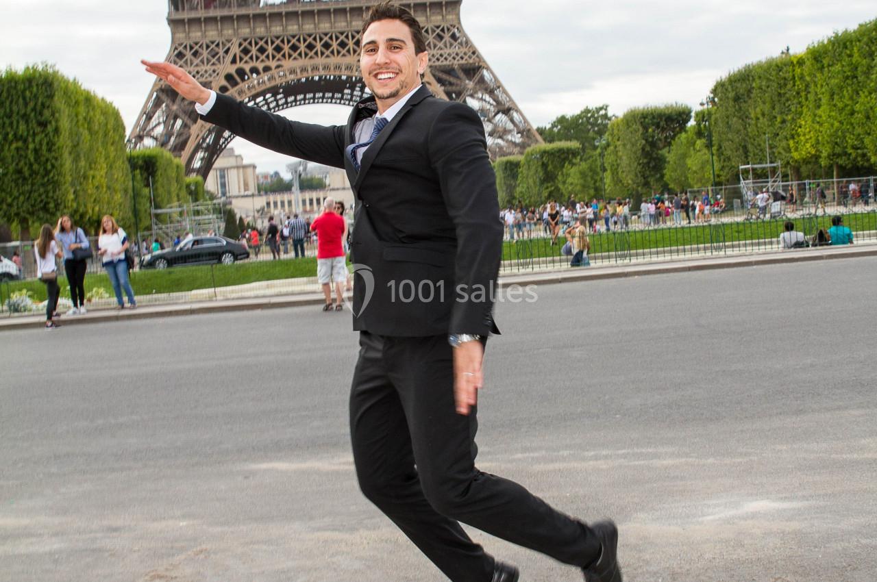 Un homme en costume souriant pose en mouvement devant la tour Eiffel, entouré de visiteurs et d'espaces verts.