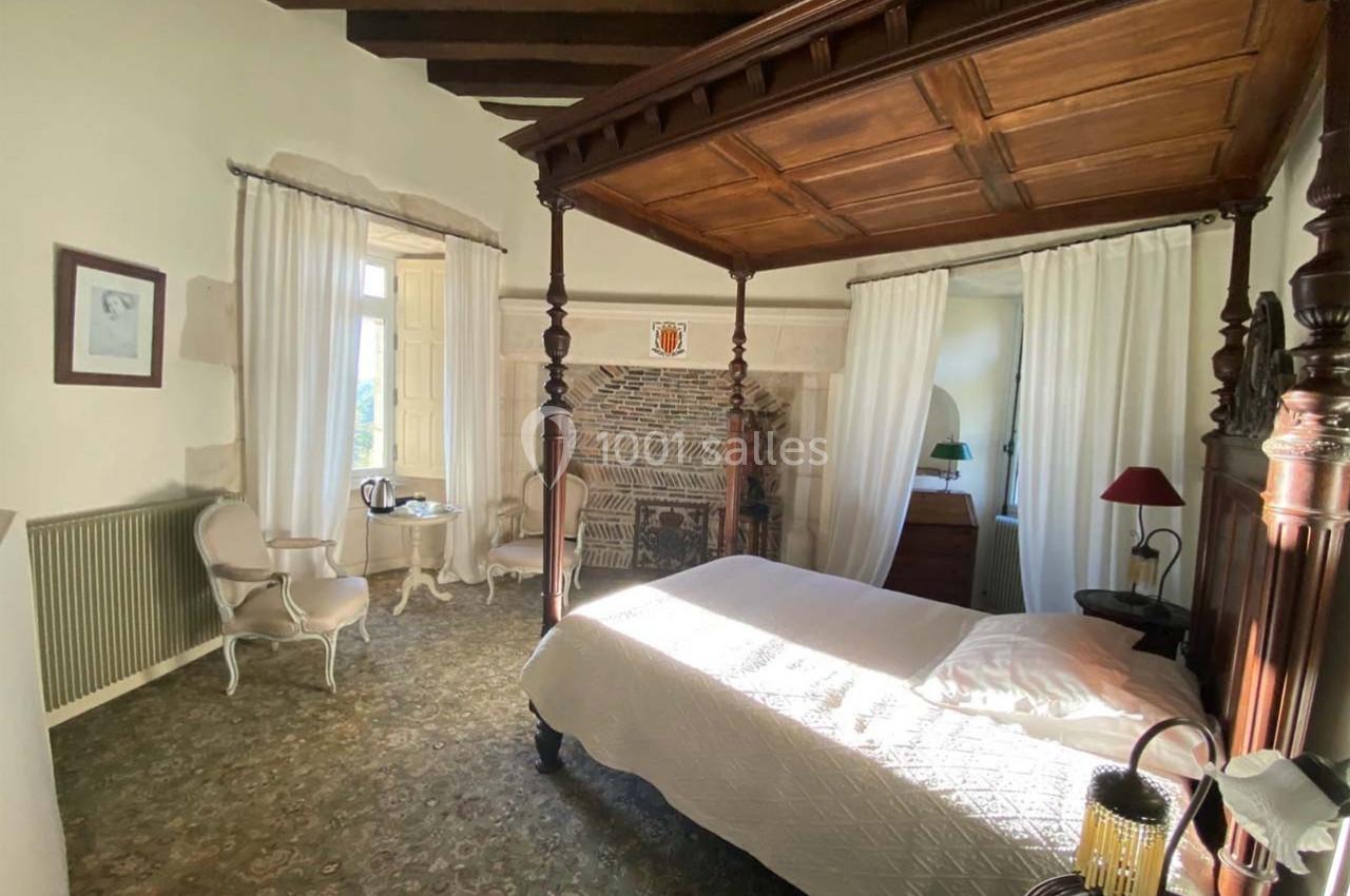 Chambre lumineuse avec lit à baldaquin en bois, cheminée en pierre, fauteuils et table près d'une fenêtre ouverte.